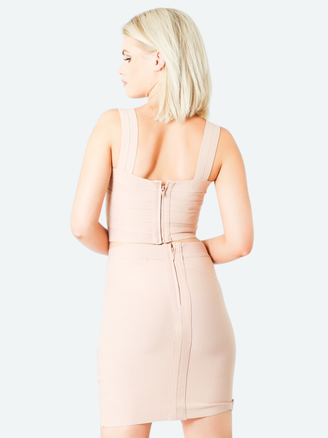 Model wears a bandage crop top with  a criss cross design, stretch fit and zip back fastening in nude colour. The model wears a matching bandage skirt to complete the look. The model has blonde hair and stands with her back to the camera. he back of the outfit can be seen, including the zip back fastening of the top and skirt.