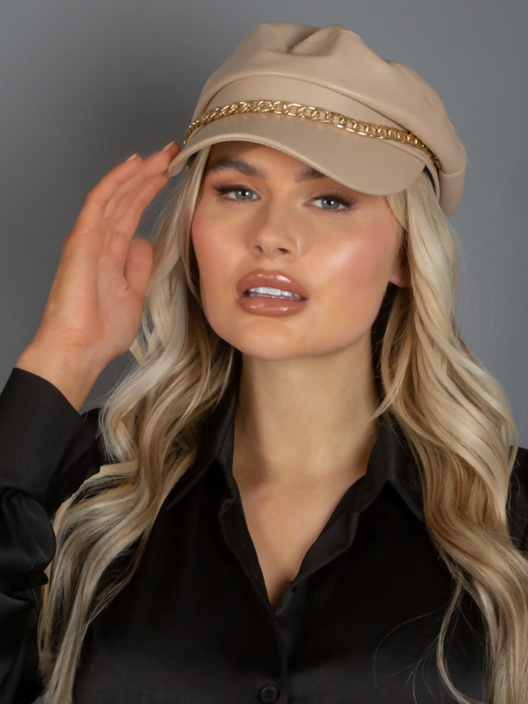 Model wears a beige pu leather paper boy hat with gold chain. The model faces the camera and places one hand to her head.