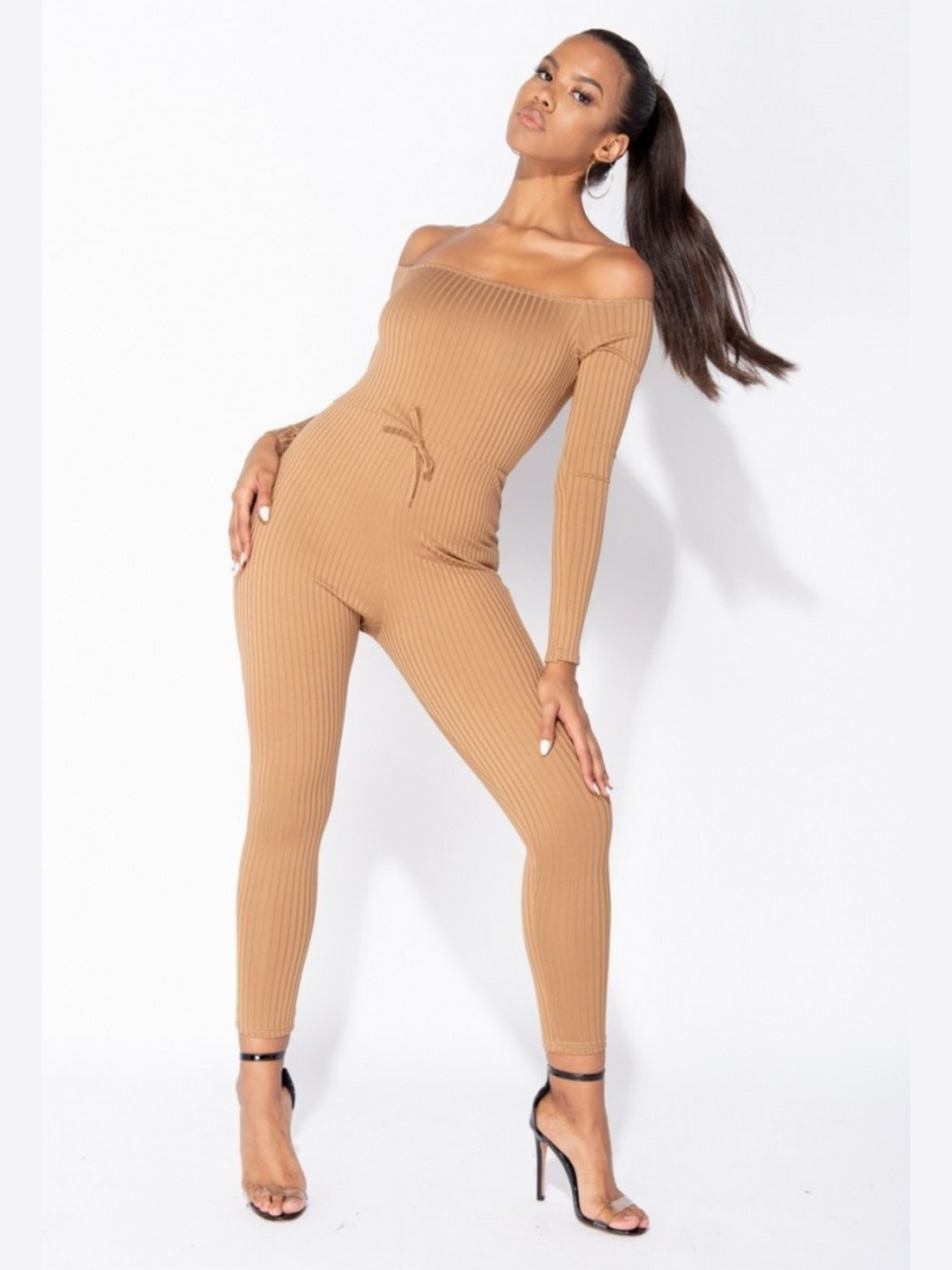 Model wears camel coloured ribbed jumpsuit with bardot neckline. Model faces the camera with one leg bent, one hand on her leg and the other on her hip. Model tilts her head, her long pony tail showing.