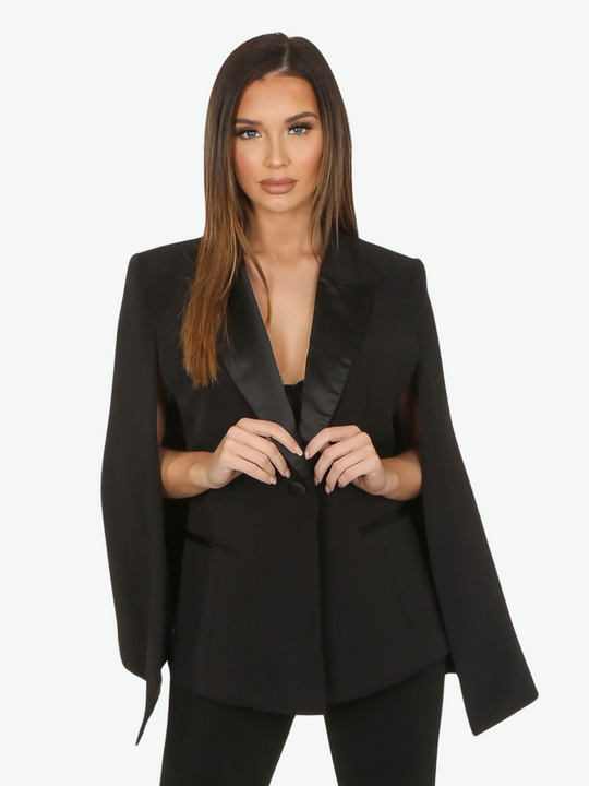 Model wears black cape blazer with side split and black satin lapel. The blazer has front button fastening.  Model places both her hands in front, touching the black satin lapel.