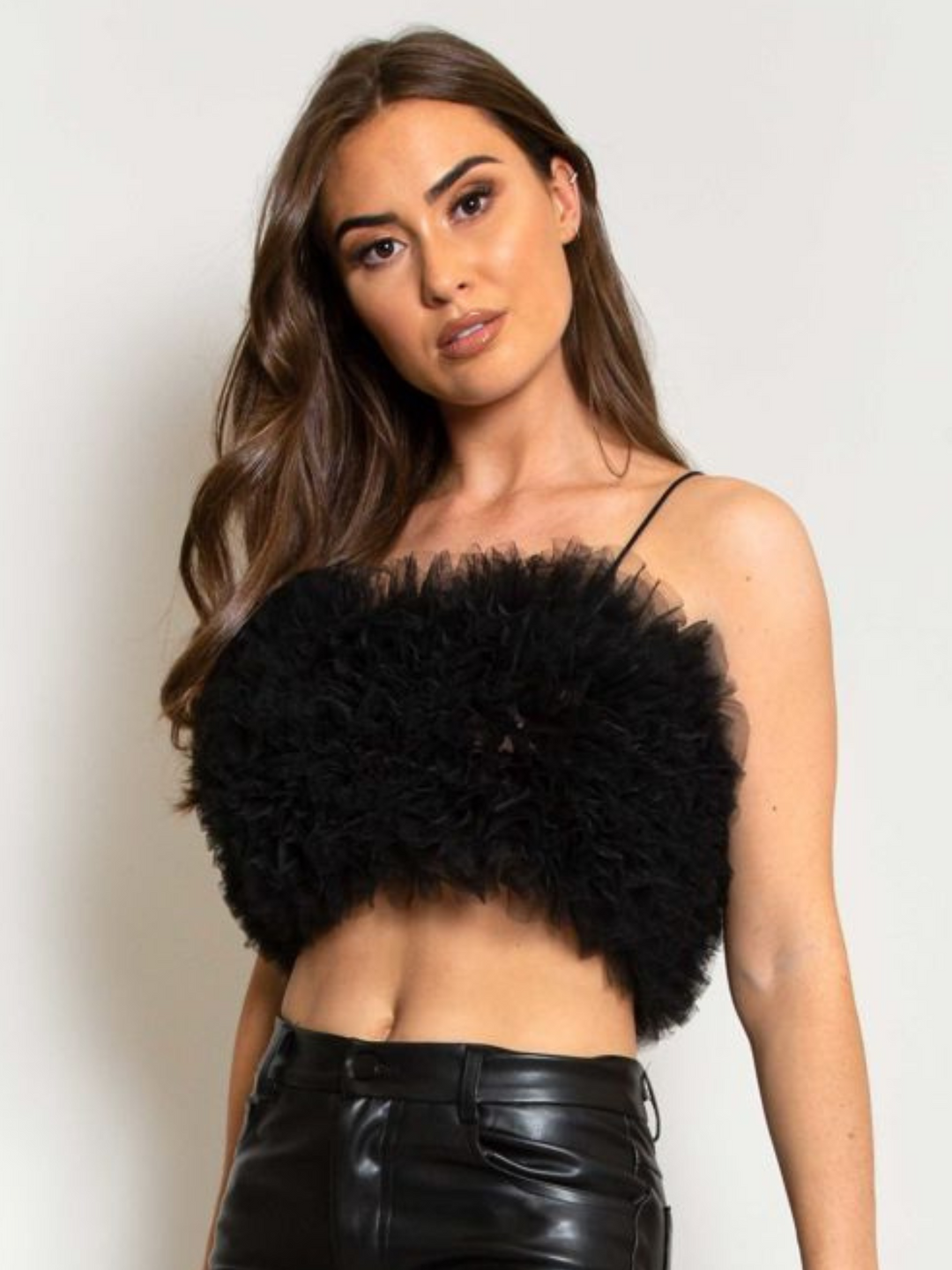 Model wears  a black organza ruffled crop top ,with spaghetti strap and zip back fastening. Model has tanned skin and long brown hair. Model faces the camera, tilting her head slightly to the side.