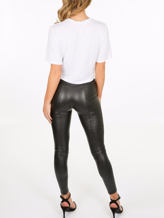 Whole body shot. Model wears a wet look, leather looking leggings with panelling. This faux leather leggings has a bodycon slim fit.  Model stands with her back to the camera with one hand in front and the other behind her back. The back of the panelled wet look leggings can be seen.