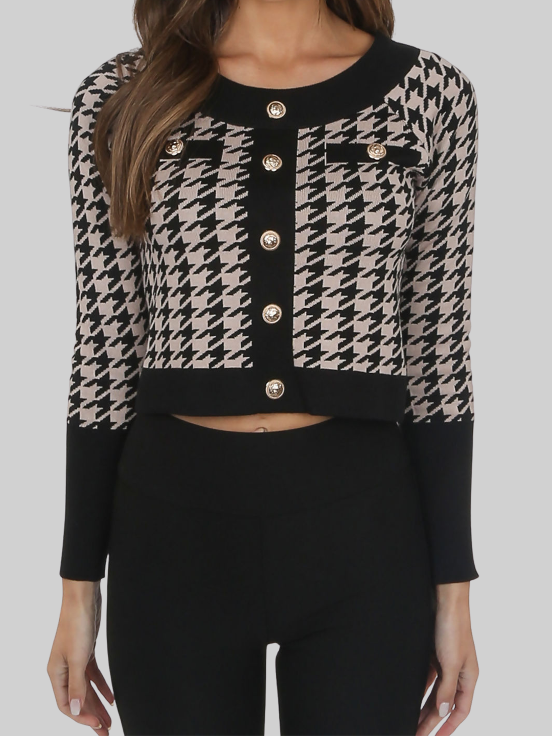 Close up. Female model stands in front of a pale grey background, her torso can be  seen.  The model wears a black and beige cropped cardigan with front military button fastening, she places both hands by her sides, the cropped cardigan can be seen.