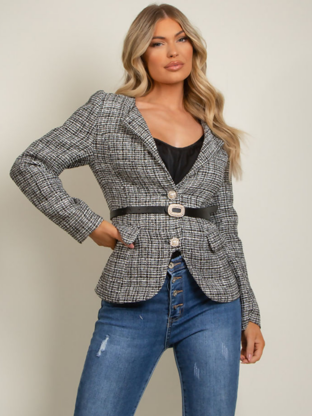 Model wears black and white tweed blazer. The glamorous blazer has diamante pearl front button fastening. The tailored blazer has an accompanying waist belt that has a diamante buckle. Model stands with one hand by her side and the other on her hip.