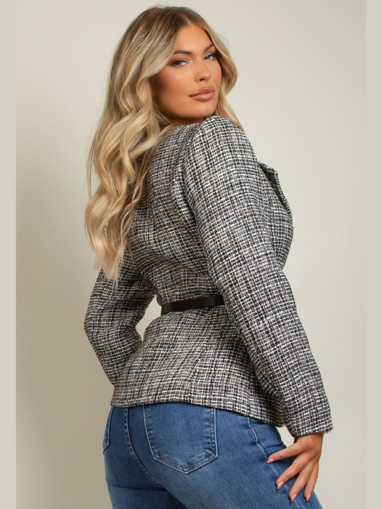 Model wears black and white tweed blazer. The glamorous blazer has diamante pearl front button fastening. The tailored blazer has an accompanying waist belt that has a diamante buckle.  Model stands to her side and turns her head to look into the camera. The back of the tweed blazer can be seen, including the black waist belt.