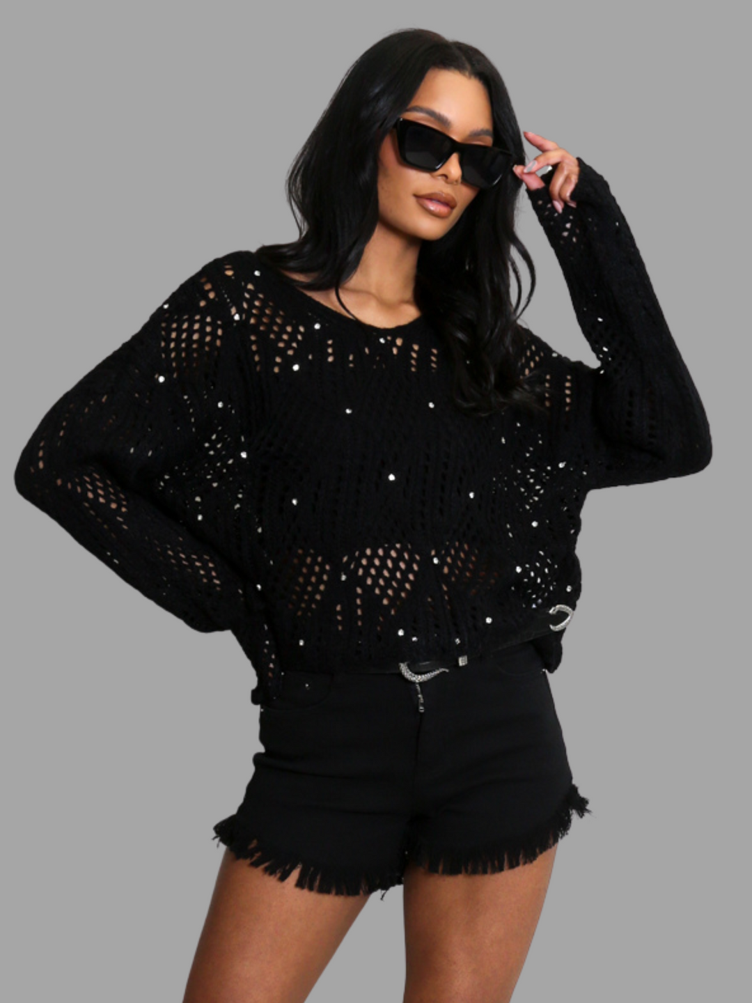 Female model wears a black crochet jumper with long sleeves and front diamante studs. The crochet jumpers has netted pattern with holes visible, the model wears a crop top underneath. The model has light brown skin and black hair, she wears sunglasses, she places one hand on her hip and the other by her head.