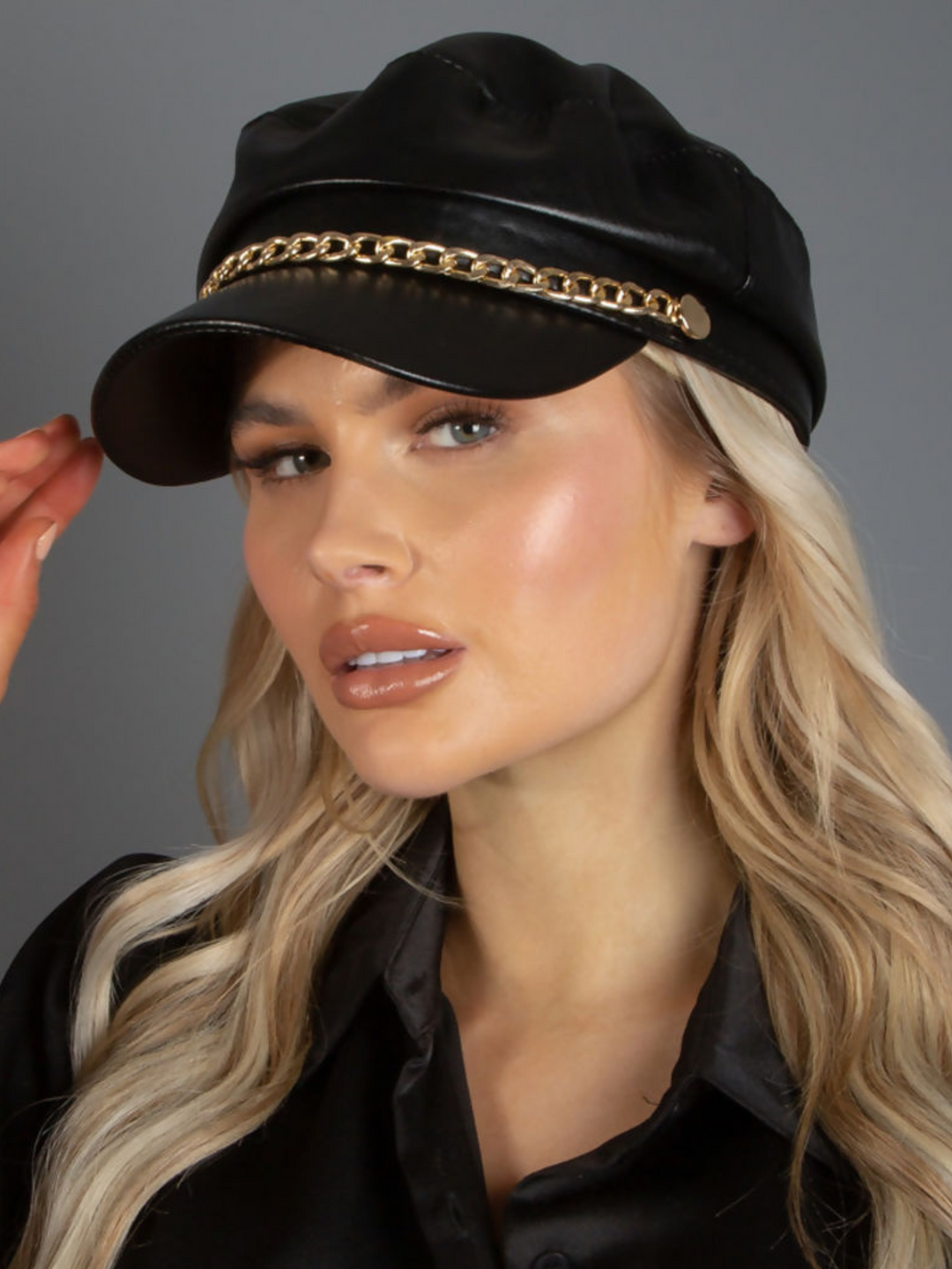 Model wears a faux leather baker boy hat with gold chain detailing at the front. Model places her hand to the pu leather cap and looks into the camera.