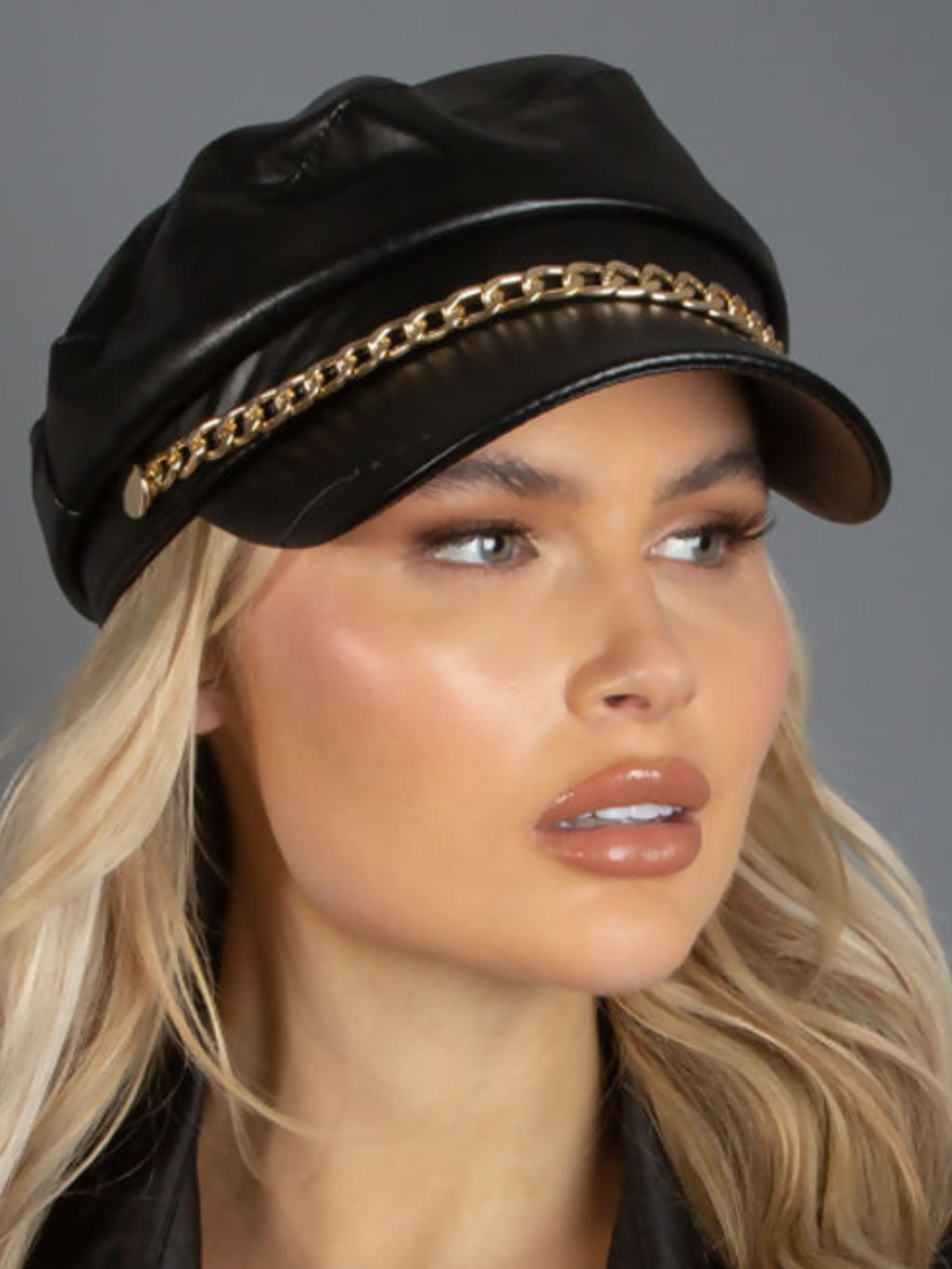 Model wears a faux leather baker boy hat with gold chain detailing at the front. Model looks off into the distance, the front of the cap can be seen.