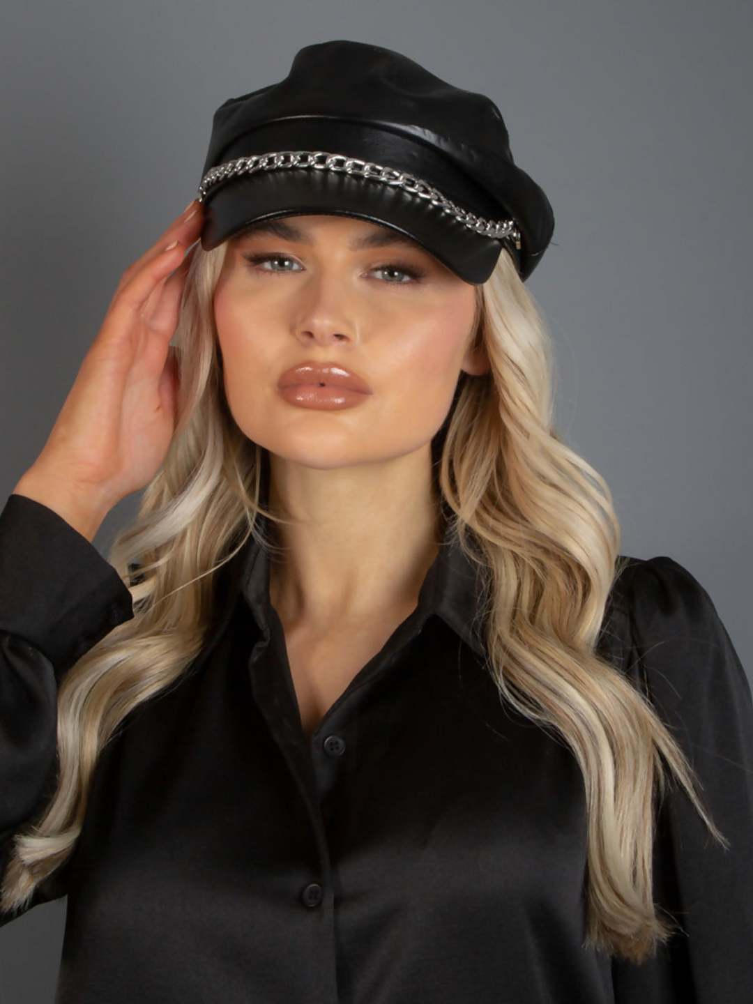 Model wears PU leather black baker boy hat with a silver chain detailing at the front. Model faces the camera and places one hand to the side of the hat.