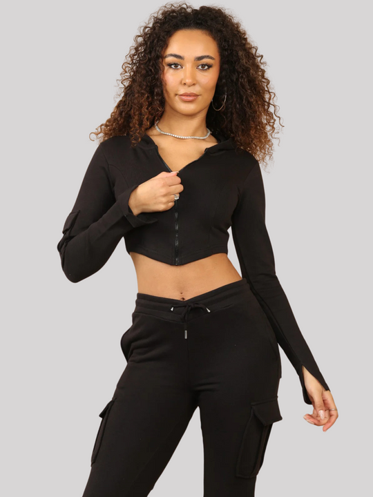Model wears a Black Cropped Top Hoodie With Zip Front Fastening and Fitted Chargo Jogger Set. The cargo jogger has pockets and one pocket on the long sleeve hoodie. Model has light brown skin, curly textured hair, she stares into the camera, placing one hand by her side and touches the cropped hoodie zip.