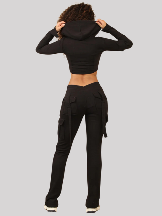 Full length shot. Model wears a Black Cropped Top Hoodie With Zip Front Fastening and Fitted Chargo Jogger Set. The cargo jogger has pockets and one pocket on the long sleeve hoodie. Model has light brown skin, curly textured hair, she stands with her back to the camera, she holds either side of the hoodie.