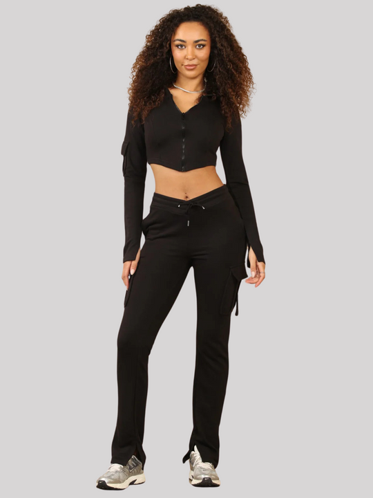 Full length shot. Model wears a Black Cropped Top Hoodie With Zip Front Fastening and Fitted Chargo Jogger Set. The cargo jogger has pockets and one pocket on the long sleeve hoodie. Model has light brown skin, curly textured hair, model stands with both hands by her side.