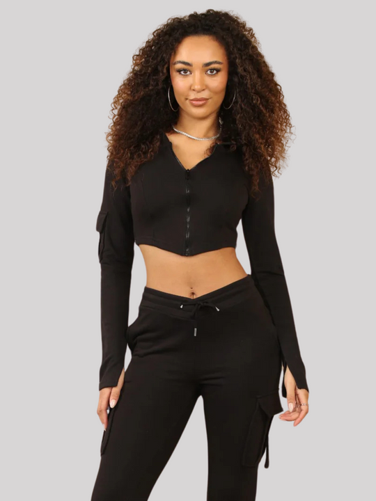 Model wears a Black Cropped Top Hoodie With Zip Front Fastening and Fitted Chargo Jogger Set. The cargo jogger has pockets and one pocket on the long sleeve hoodie. Model has light brown skin, curly textured hair, model faces the camera, she stands with her hands by her side.
