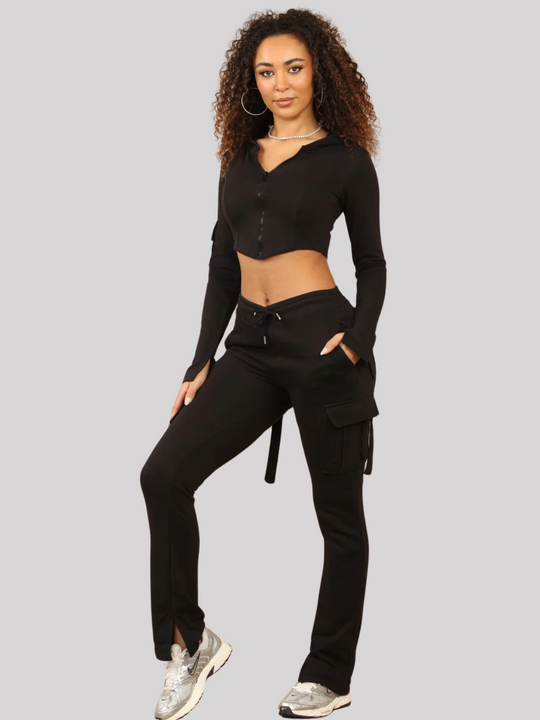 Full length shot. Model wears a Black Cropped Top Hoodie With Zip Front Fastening and Fitted Chargo Jogger Set. The cargo jogger has pockets and one pocket on the long sleeve hoodie. Model has light brown skin, curly textured hair. Model stands with one hand in his pocket and he other on her leg, she stands slightly to the side.