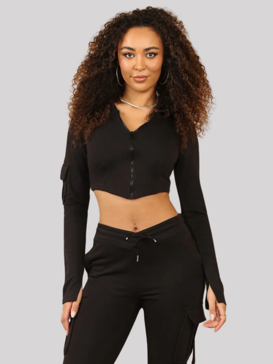 Model wears a Black Cropped Top Hoodie With Zip Front Fastening and Fitted Chargo Jogger Set. The cargo jogger has pockets and one pocket on the long sleeve hoodie. Model has light brown skin, curly textured hair, model faces the camera, she stands with her hands by her side.