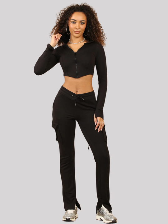 Full length shot. Model wears a Black Cropped Top Hoodie With Zip Front Fastening and Fitted Chargo Jogger Set. The cargo jogger has pockets and one pocket on the long sleeve hoodie. Model has light brown skin, curly textured hair, she places one hand on her leg and the oth by her hair.