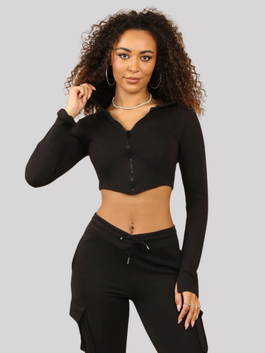 Model wears a Black Cropped Top Hoodie With Zip Front Fastening and Fitted Chargo Jogger Set. The cargo jogger has pockets and one pocket on the long sleeve hoodie. Model has light brown skin, curly textured hair, she places one hand on her leg and the other touches her hair.