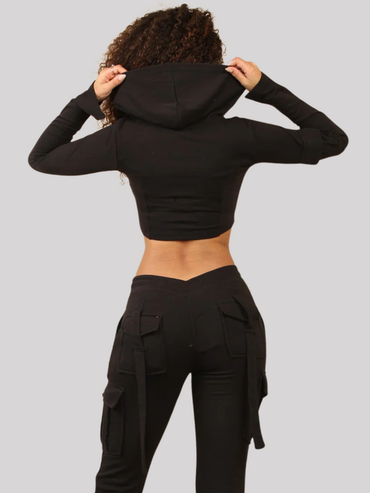 Model wears a Black Cropped Top Hoodie With Zip Front Fastening and Fitted Chargo Jogger Set. The cargo jogger has pockets and one pocket on the long sleeve hoodie. Model has light brown skin, curly textured hair, she stands with her back to the camera, the hooded crop top and back of the cargo joggers can be see.