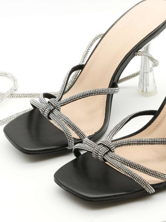 Product close up shot. The shoe is a  black patentt leather look muled shoe, with open toes and silver diamante cross over straps. The shoe also has a silver diamante strap that  fis designed to fasten along the shin.