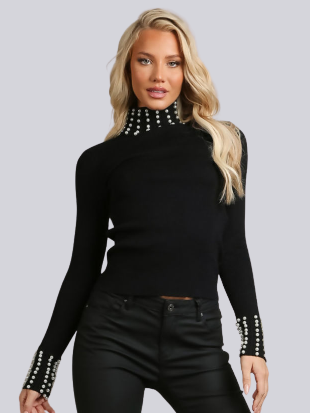 Female model wears a black knitted turtle neck jumper, with pearl  detailed pearl accents at the high neck and  cuff. The female model has tanned skin and long blonde hair, she faces the camera placing her hands by her sides.