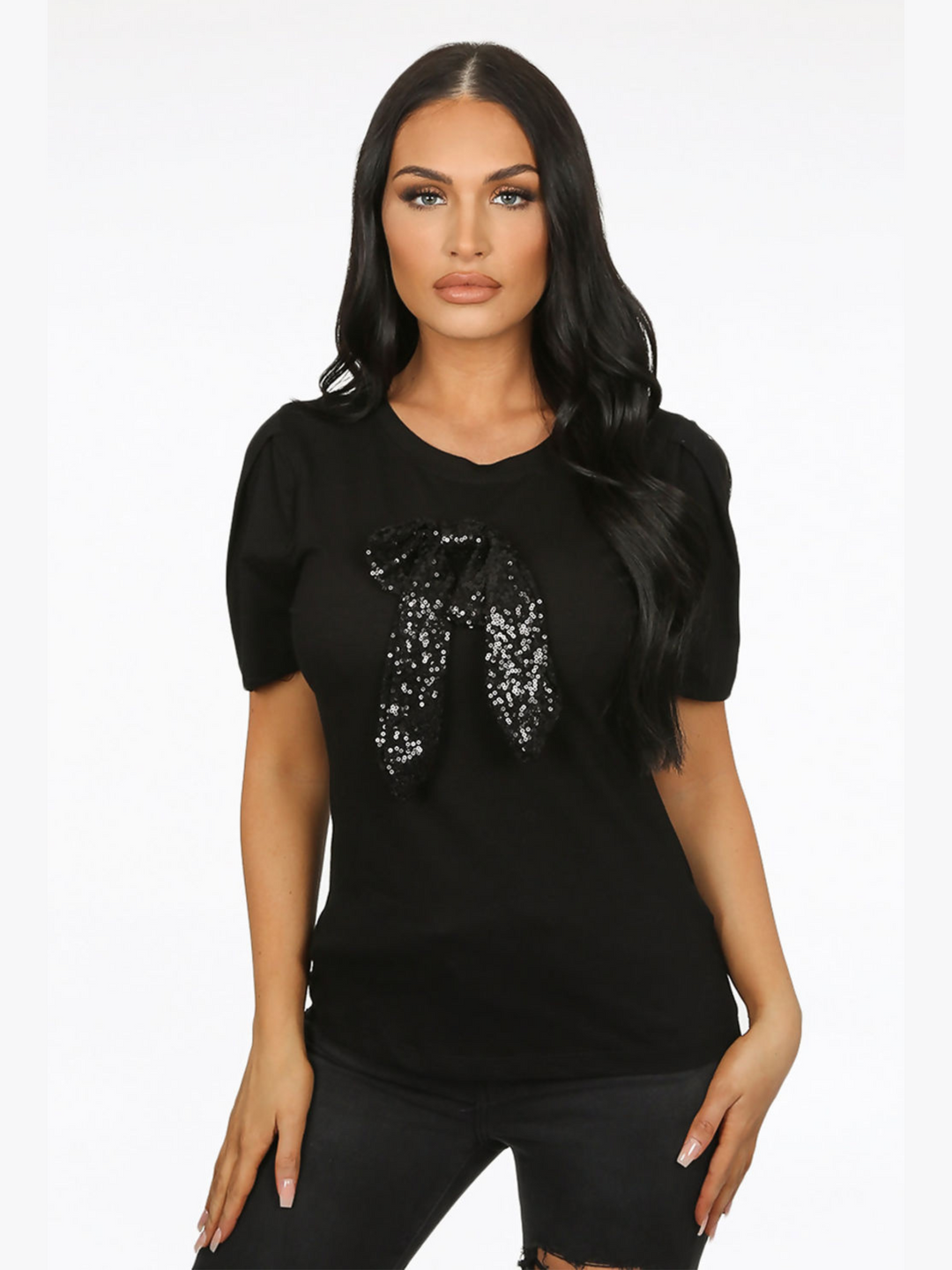 Female model wears a luxe black t shirt with a rounded neck amd 3/4 sleeves, with a large black sequin bow. The model faces the camera in front of  a plain background,  she has tanned skin and long black hair. The model places both hands by her sides.