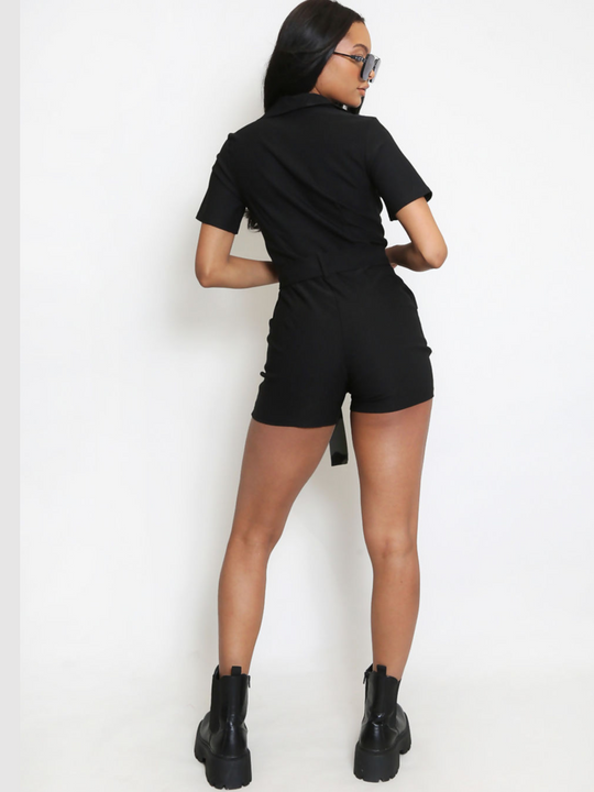Full length shot. Model wears black playsuit with exposed front silver zip, waist tie belt silver accent buttons and mid sleeved detailing. The model has brown skin and long black hair, she wears  oversized blue tinted sunglasses. Model stands with her back to the camera, the back of the playsuit can be seen, including the playsuit's waist  belt and collar.