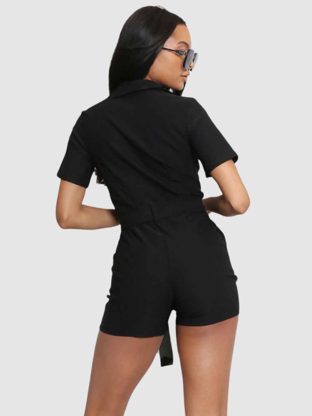 Model wears black playsuit with exposed front silver zip, waist tie belt silver accent buttons and mid sleeved detailing. The model has brown skin and long black hair, she wears  oversized blue tinted sunglasses.  Model stands with her back to the camera, the back of the plasysuit can be seen, including the front tie belt and collar.