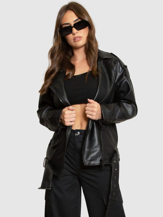 Female model stands in front of a grey background, she wears an oversized pu leather jacket , she faces the camera; she has tanned skin and long brown hair. She stands with her jacket open and places both hands on either side of the jacket, she wears black sun glasses.