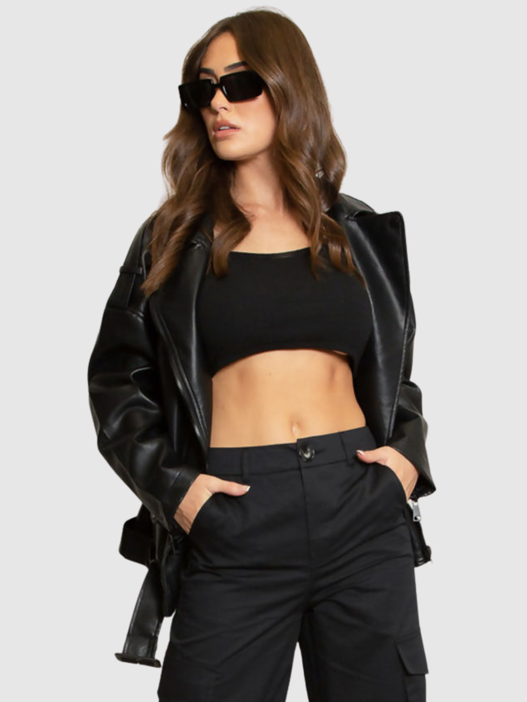 Female model stands in front of a grey background, she wears an oversized pu leather jacket , she faces the camera; she has tanned skin and long brown hair. She stands with her jacket open and places both hands in her trouser pockets..