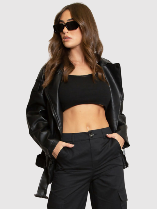 Female model stands in front of a grey background, she wears an oversized pu leather jacket , she faces the camera; she has tanned skin and long brown hair. She stands with her jacket open and places both hands in her trouser pockets..