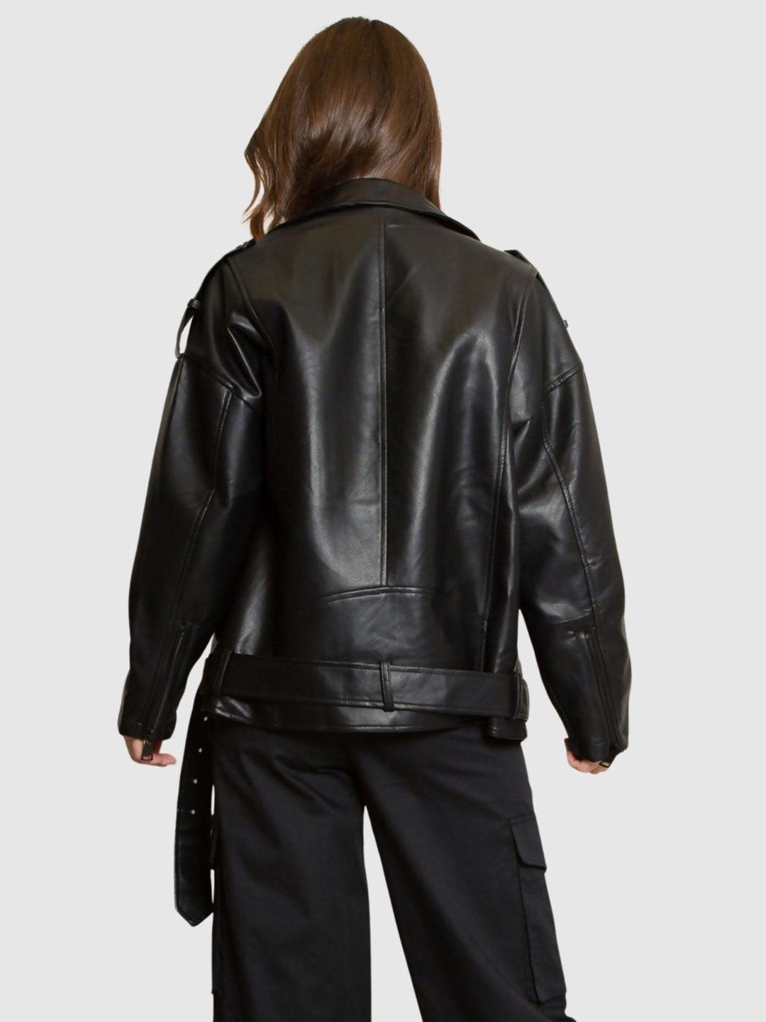 Female model stands in front of a grey background, she wears an oversized pu leather jacket , she faces her back to the camera;  the back of the  oversized pu leather jacket can be seen, including the low rise belt.