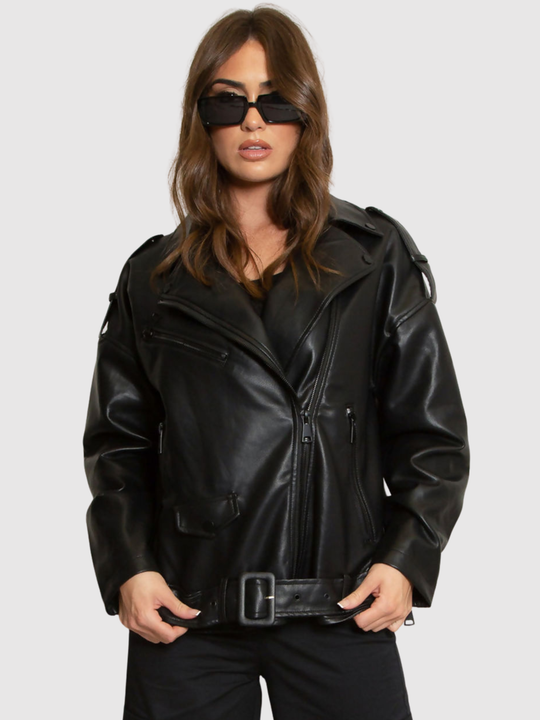 Female model stands in front of a grey background, she wears an oversized pu leather jacket , she faces the camera; she has tanned skin and long brown hair. She faces the cameran and wears the jacket closed, the oversized biker jacket style can be seen, including the low rise belt.