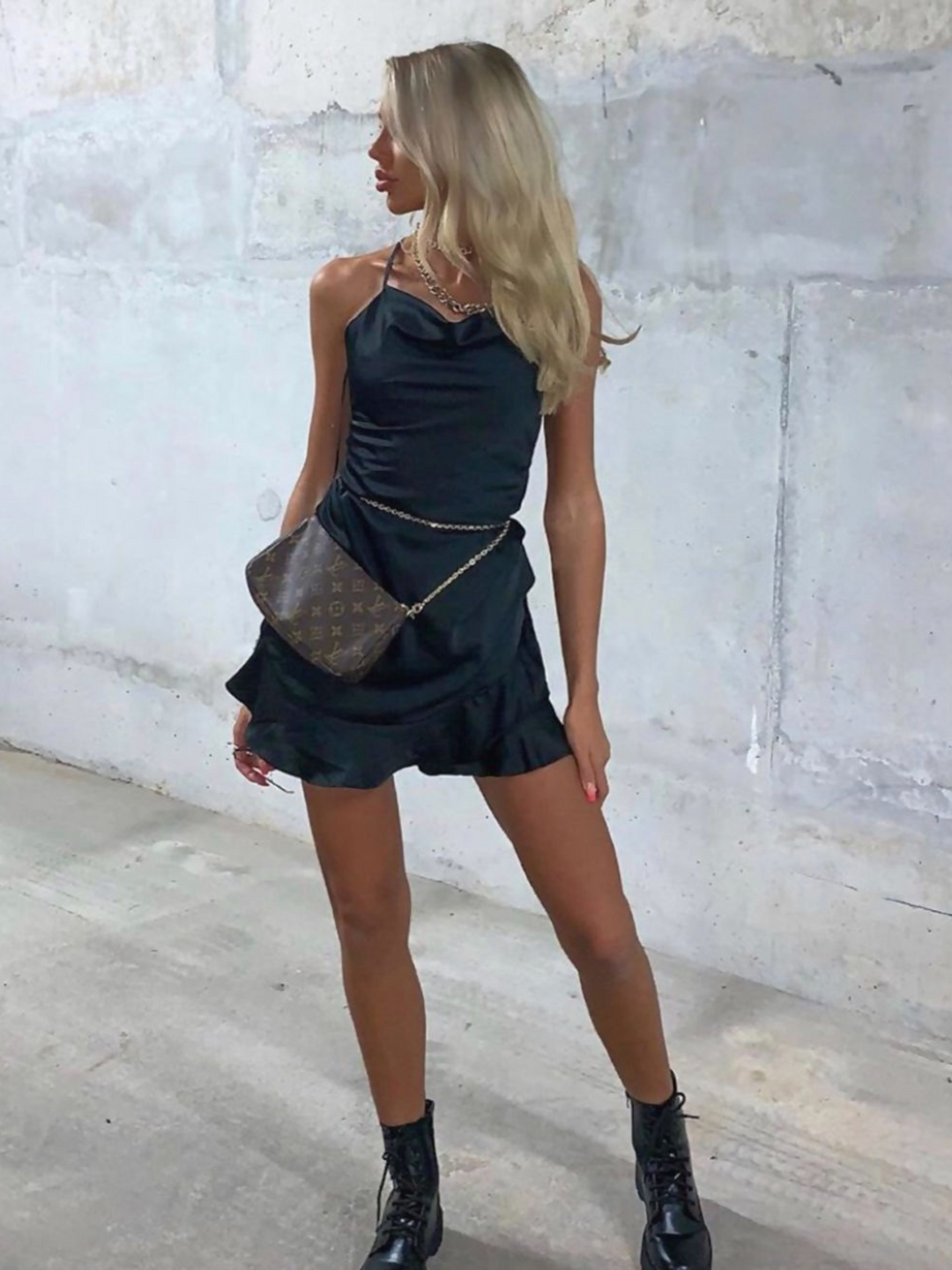 Model wears black satin playsuit, with spaghetti straps, cowl neckline and frill trimmed hem. Model wears a statement bag at her waist.