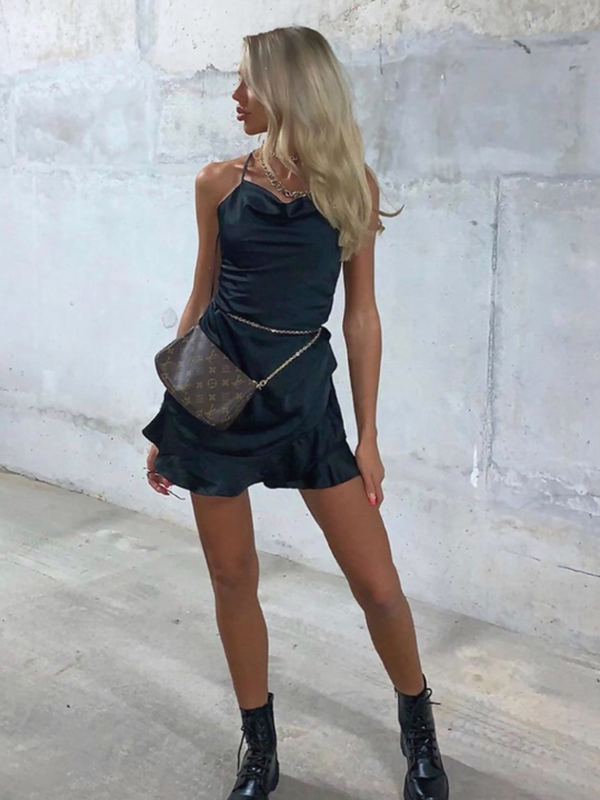Model wears black satin playsuit, with spaghetti straps, cowl neckline and frill trimmed hem. Model wears a statement bag at her waist.