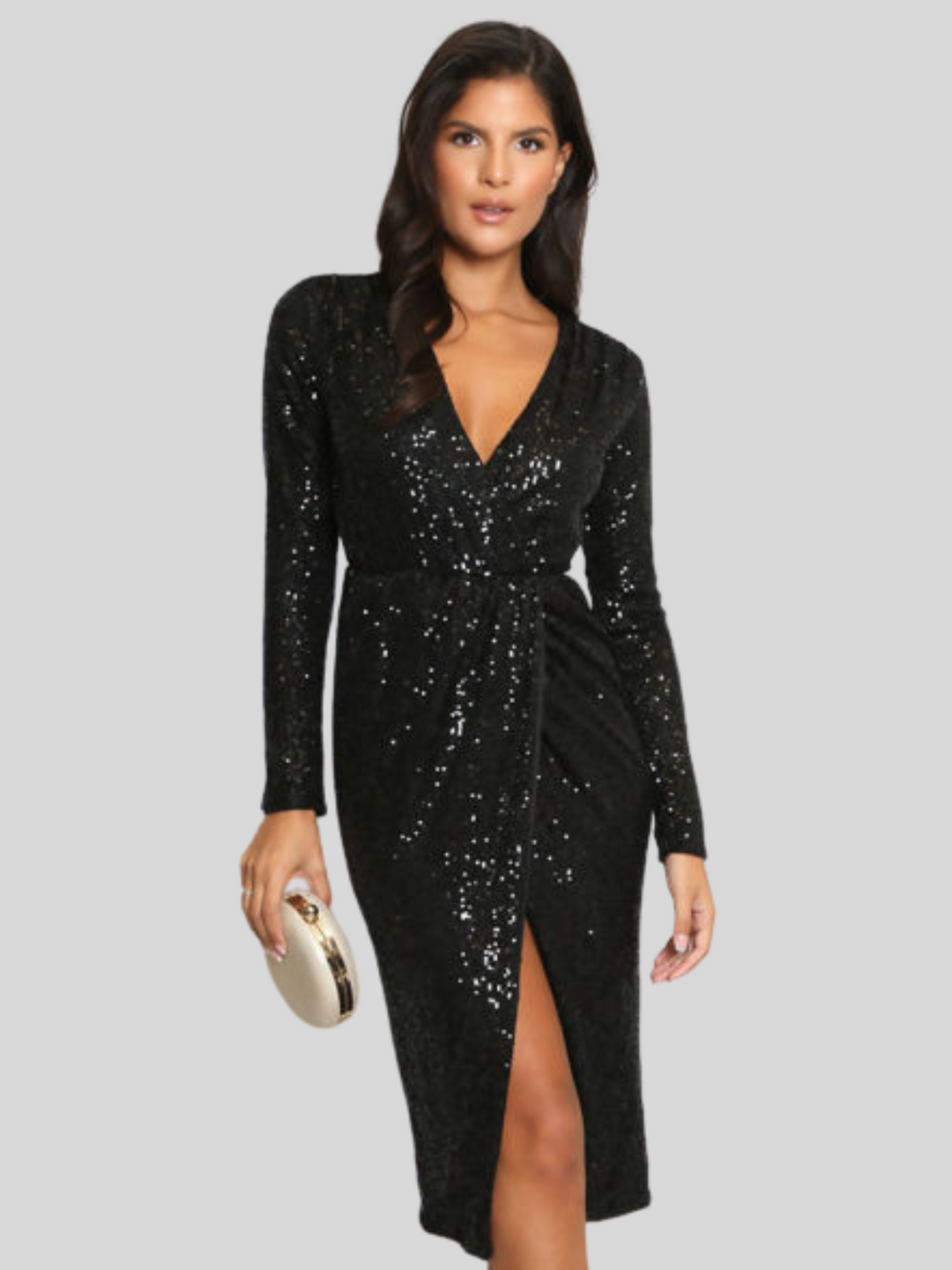 Mid length shot. Model wears a black sequin midaxi dress with a v neckline, long sleeves and a split front cross over front. The model has tanned skin and  long brown hair, she looks directly int the camera. The model holds a hand bag in one hand and places the other by her side.