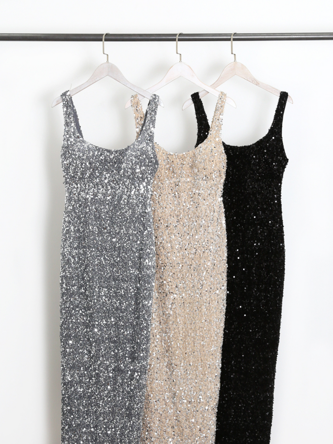 Three sequin midi dresses with  a square cut neckline hand on a clothing rack. The dresses all have the same design, the colour varies, these are: silver, cream and black.