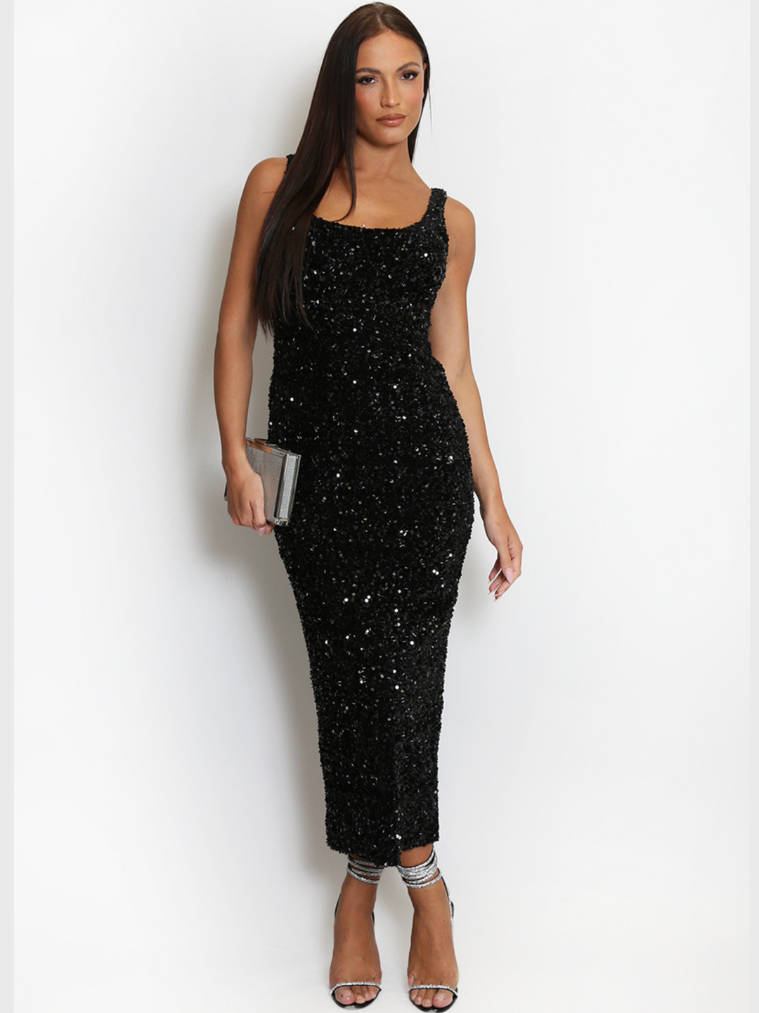 Model weas a  deluxe black sequin midi dress with a square neckline and a sleeveless cut. The model has tanned skin and long brown hair. The model faces the camera, the front of the sequin dress can be seen, she wears a strappy diamante heel and crosses over her feet, she holds a silver clutch bag in one hand and her hand by her side.