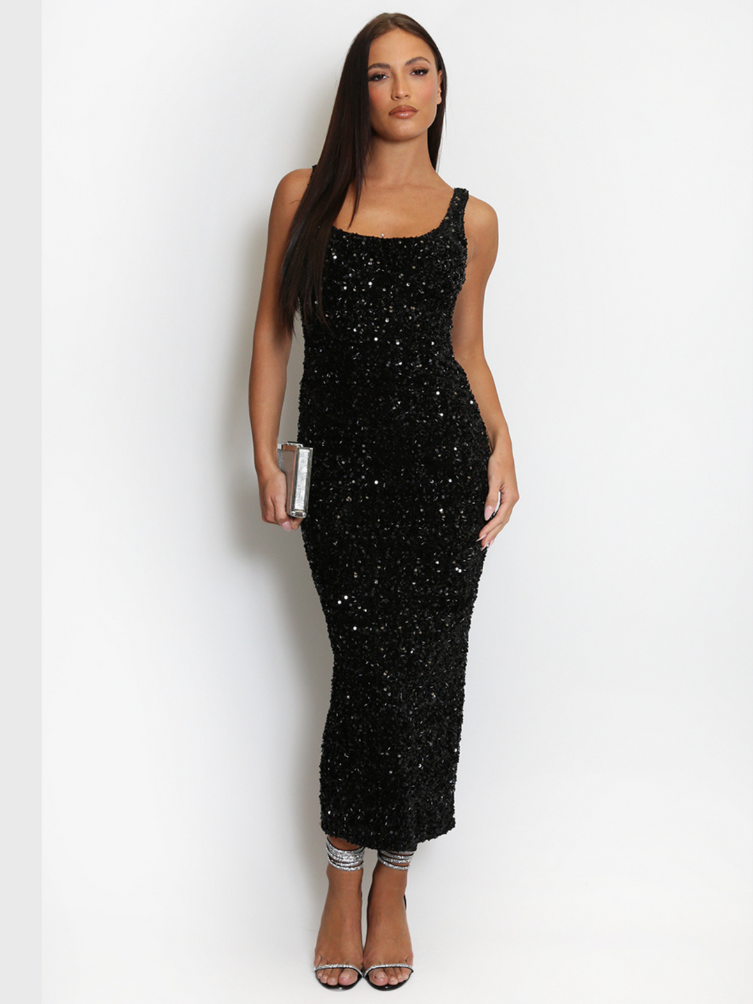 Full length shot. Model weas a  deluxe black sequin midi dress with a square neckline and a sleeveless cut. The model has tanned skin and long brown hair. The model faces the camera, she places one hands by her side and holds a silver clutch in the other.