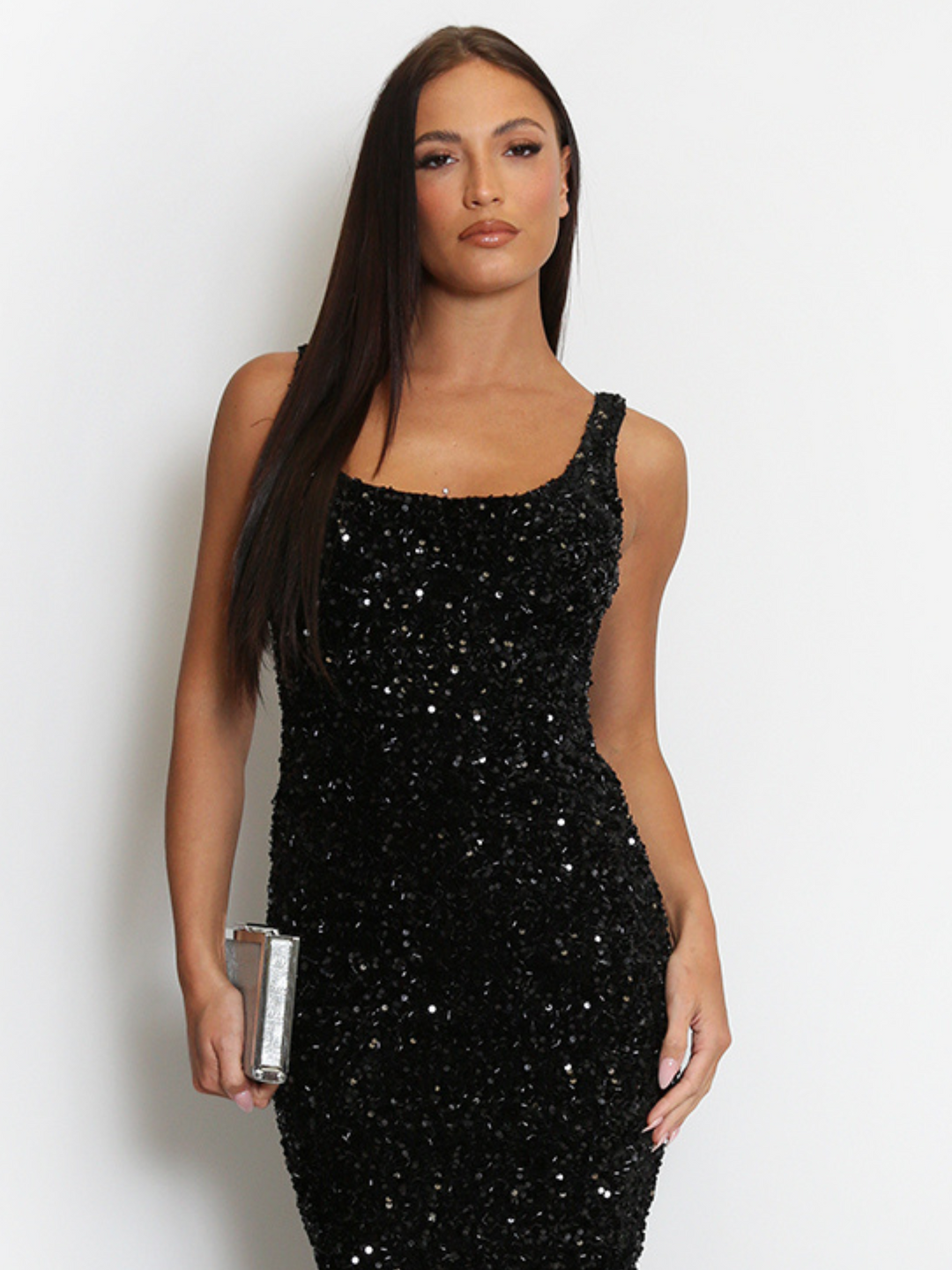 Mid length shot. Model weas a  deluxe black sequin midi dress with a square neckline and a sleeveless cut. The model has tanned skin and long brown hair. The model faces forward, holding a silver clutch in one hand and places her hand by her side with the other.