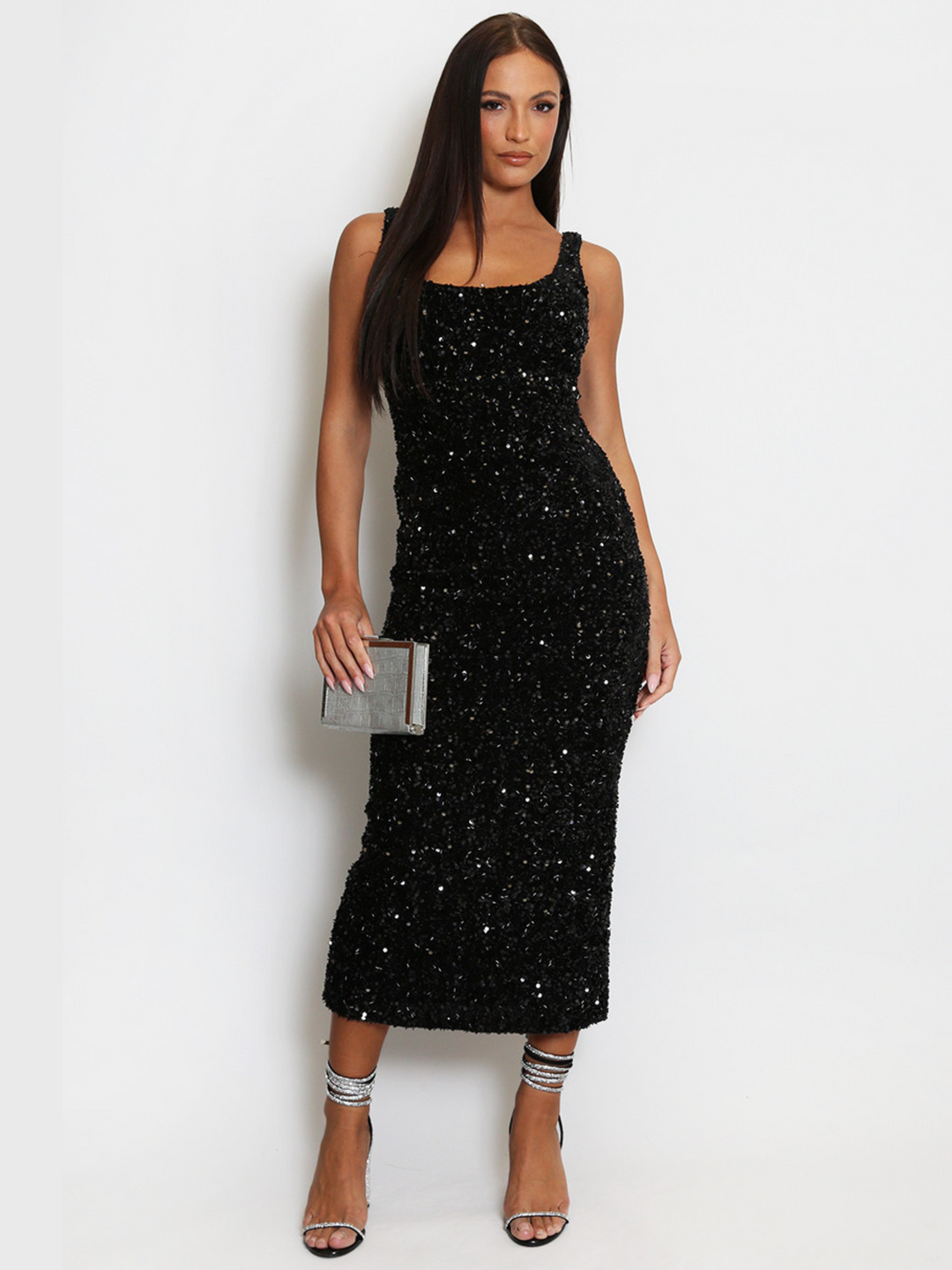 Full length shot. Model weas a  deluxe black sequin midi dress with a square neckline and a sleeveless cut. The model has tanned skin and long brown hair. The model faces the camera, holding a silver clutch bag  and strappy diamante lace  up heels.