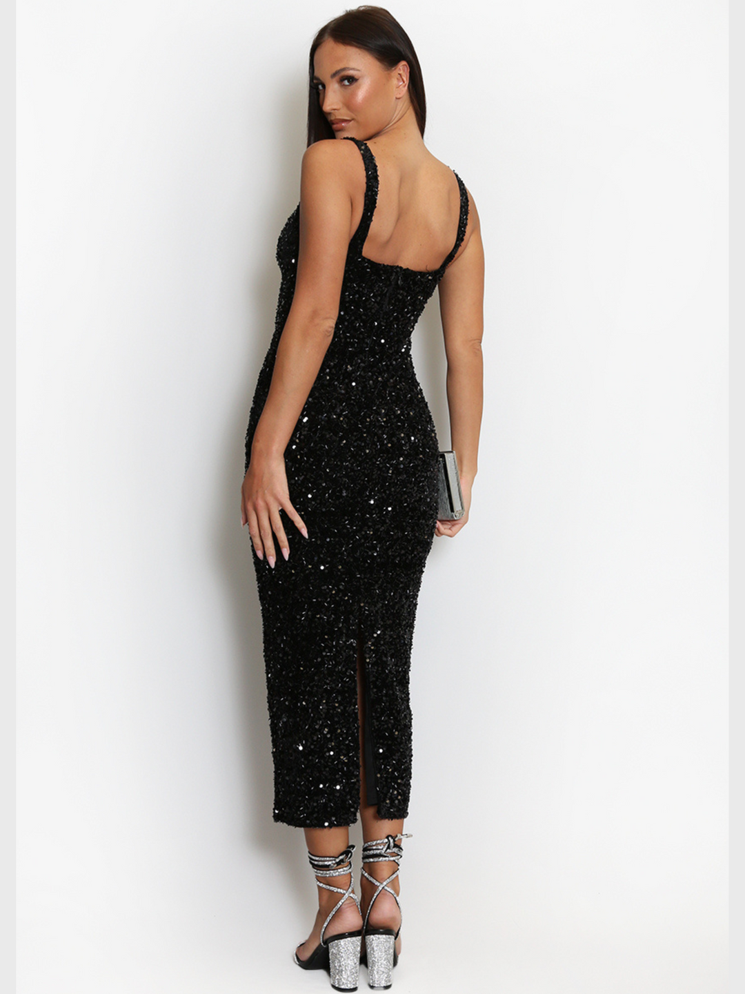 Full length shot. The model stands with her back to the camera, the back of the dress can be seen; including the square cut black design just below the shoulder blade, the long back of the dress that has a slit and the back of the diamante lace up shoe.