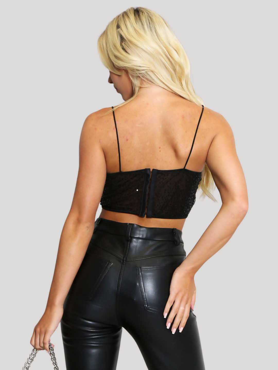 Female model stands before a plain backround. The model wears a  black sequin crop top with hook back fastening and spaghetti straps. The crop top has a front sheer panel with sequin detailing. The model has tanned skin and long blonde hair, she stands with her back  to the camera, the hood bak fastening and the back of the pu leather trousers can be seen.