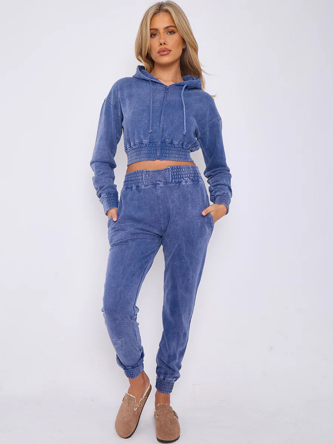 Full length shot. Model wears a blue acid wash zip up hoodie cropped  top that has an elasticated waist. The blue acid wash joggers has a matchig joggers set, that has an elasticated waist and side pockets. Model has long blonde hair, she faces the camera and stads with her hands in her pockets.