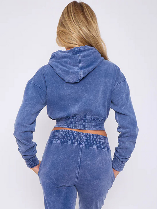 Model wears a blue acid wash zip up hoodie cropped  top that has an elasticated waist. The blue acid wash joggers has a matchig joggers set, that has an elasticated waist and side pockets. Model has long blonde hair, she stands with her back to the camera, the back of the cropped top and hoodie can be seen. The model places her hands in her pockets, the acid wash hood can also be seen.