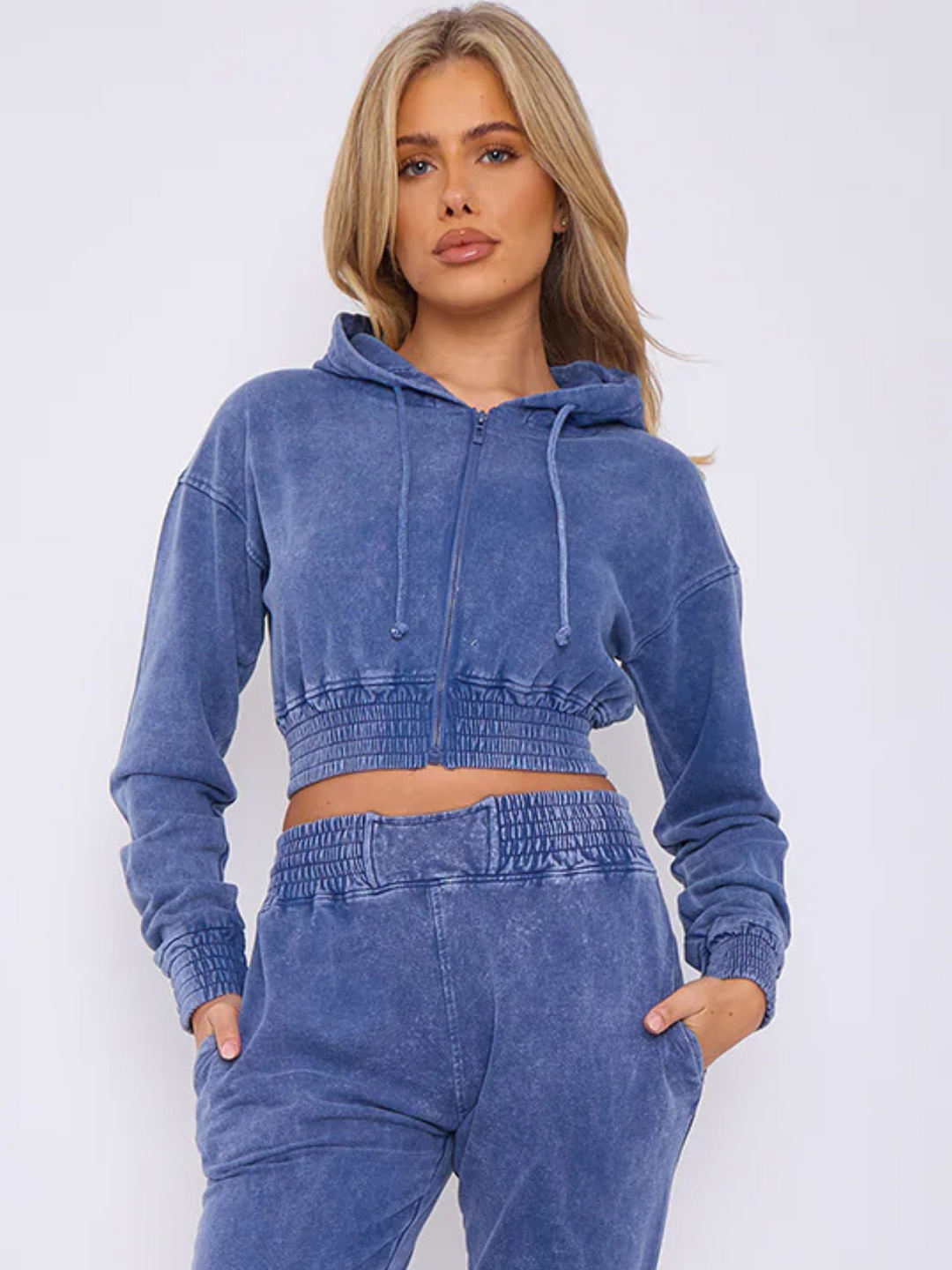 Model wears a blue acid wash zip up hoodie cropped  top that has an elasticated waist. The blue acid wash joggers has a matchig joggers set, that has an elasticated waist and side pockets. Model has long blonde hair, she faces the camera and places her hans in her pockets. The front of the draw string hoodie can be seen.