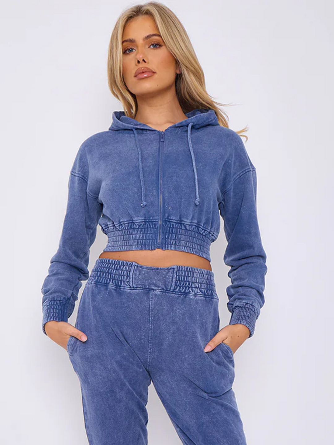 Model wears a blue acid wash zip up hoodie cropped  top that has an elasticated waist. The blue acid wash joggers has a matchig joggers set, that has an elasticated waist and side pockets. Model has long blonde hair, she places her hands in her pockets.