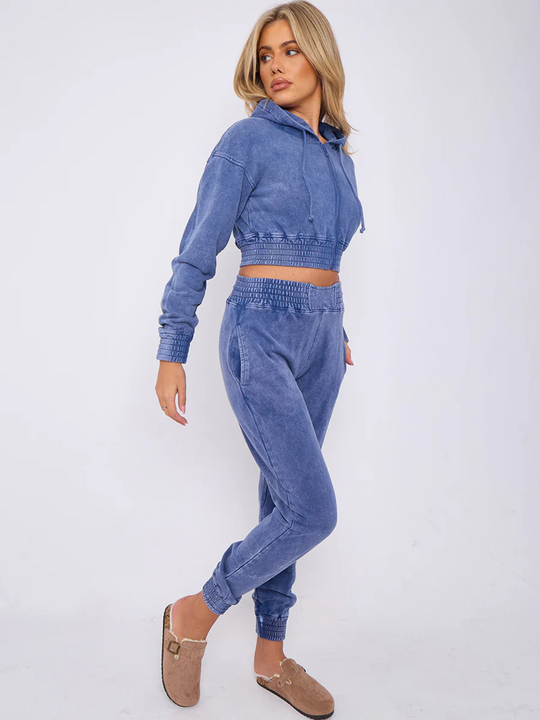 Full length shot. Model wears a blue acid wash zip up hoodie cropped  top that has an elasticated waist. The blue acid wash joggers has a matchig joggers set, that has an elasticated waist and side pockets. Model has long blonde hair, she stands to the side and turns her head to look behind her. She has one hand in her pocket and the other by her side. The side of the joggers set can be seen.