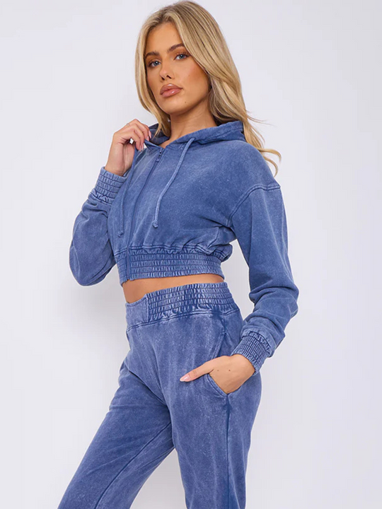 Model wears a blue acid wash zip up hoodie cropped  top that has an elasticated waist. The blue acid wash joggers has a matchig joggers set, that has an elasticated waist and side pockets. Model has long blonde hair, she stands to the side and places her hand in her pocket, she turns her head to the side and looks into the camera.