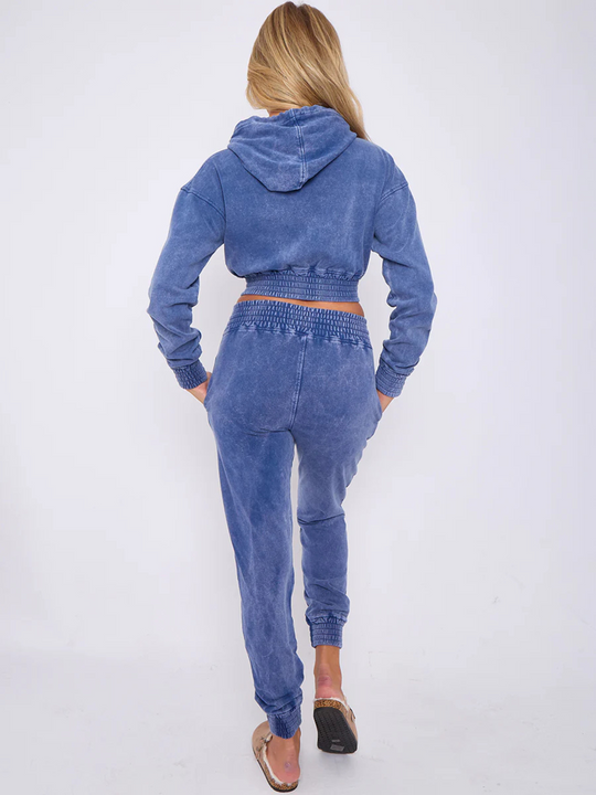 Full length shot. Model wears a blue acid wash zip up hoodie cropped  top that has an elasticated waist. The blue acid wash joggers has a matchig joggers set, that has an elasticated waist and side pockets. Model has long blonde hair, she stands with her back to the camera, placing both hands into the side pockets, the hood is also visible.