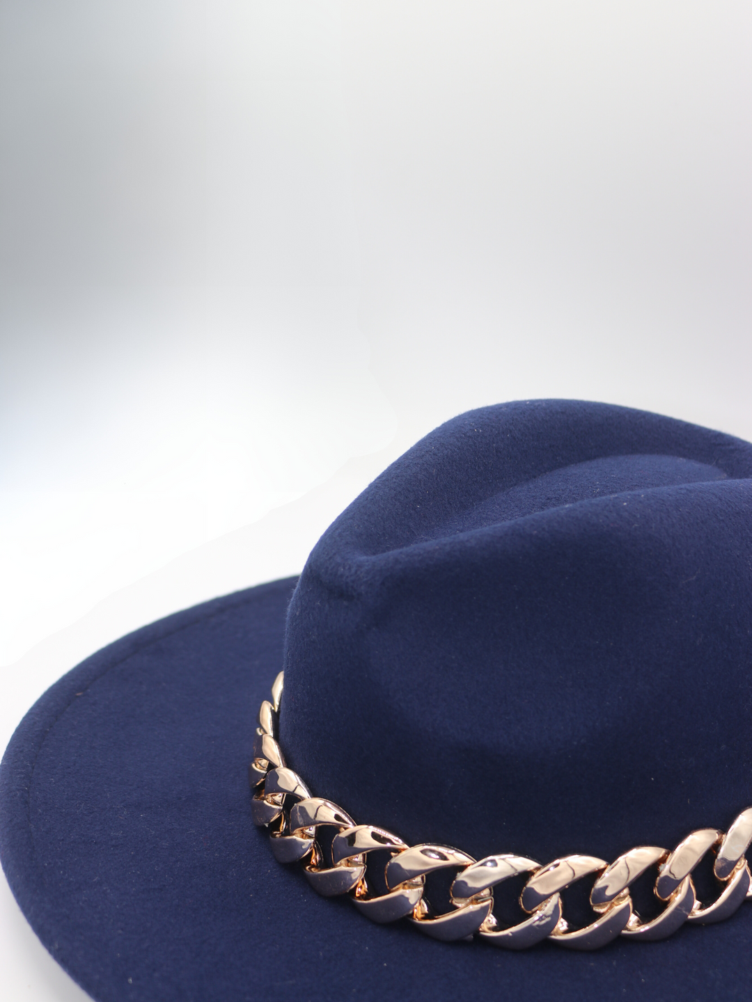 The  side of the navy felt fedora hat  with gold linked chunky chain detailing can be seen.  The top of the hat is also visible.